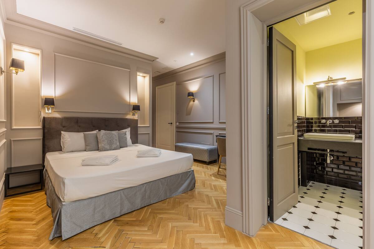 Habitación de hotel con una cama grande, un banco, un baño con piso de baldosas e iluminación empotrada.