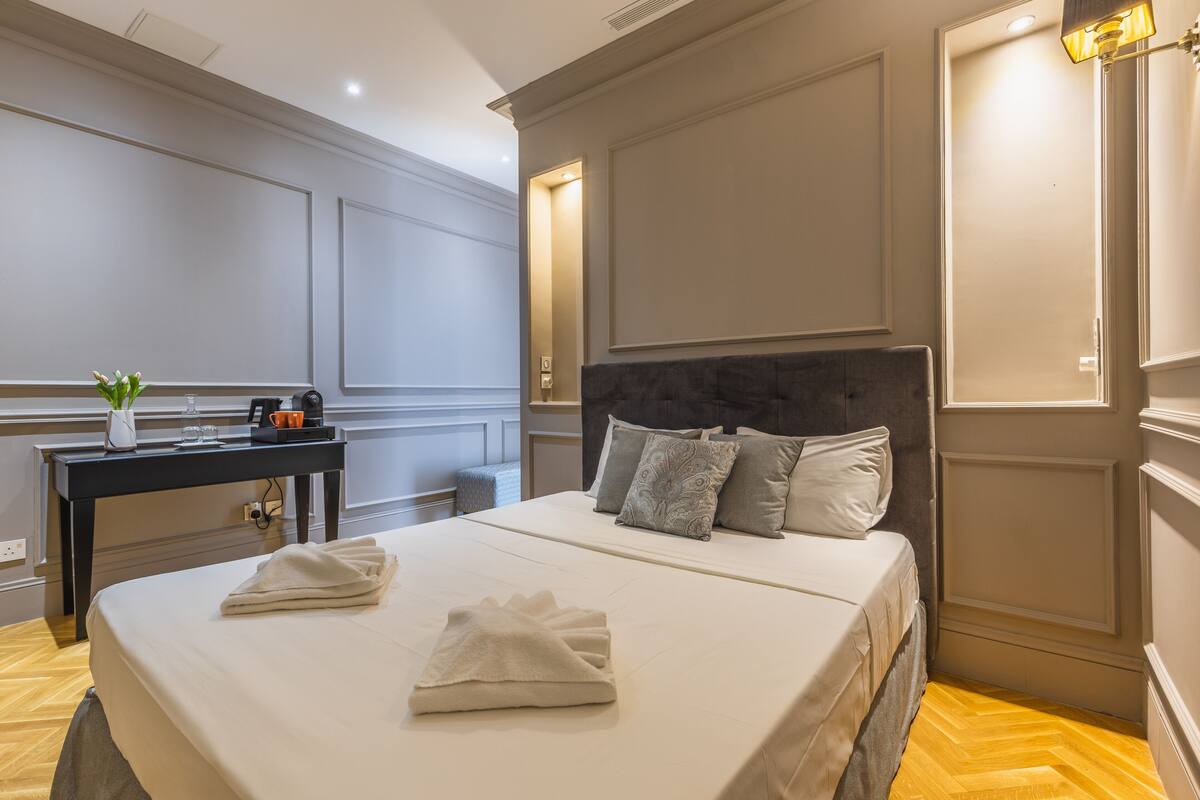 Una habitación de hotel con una cama bien hecha, una mesita de noche con lámpara y un pequeño escritorio con una planta.
