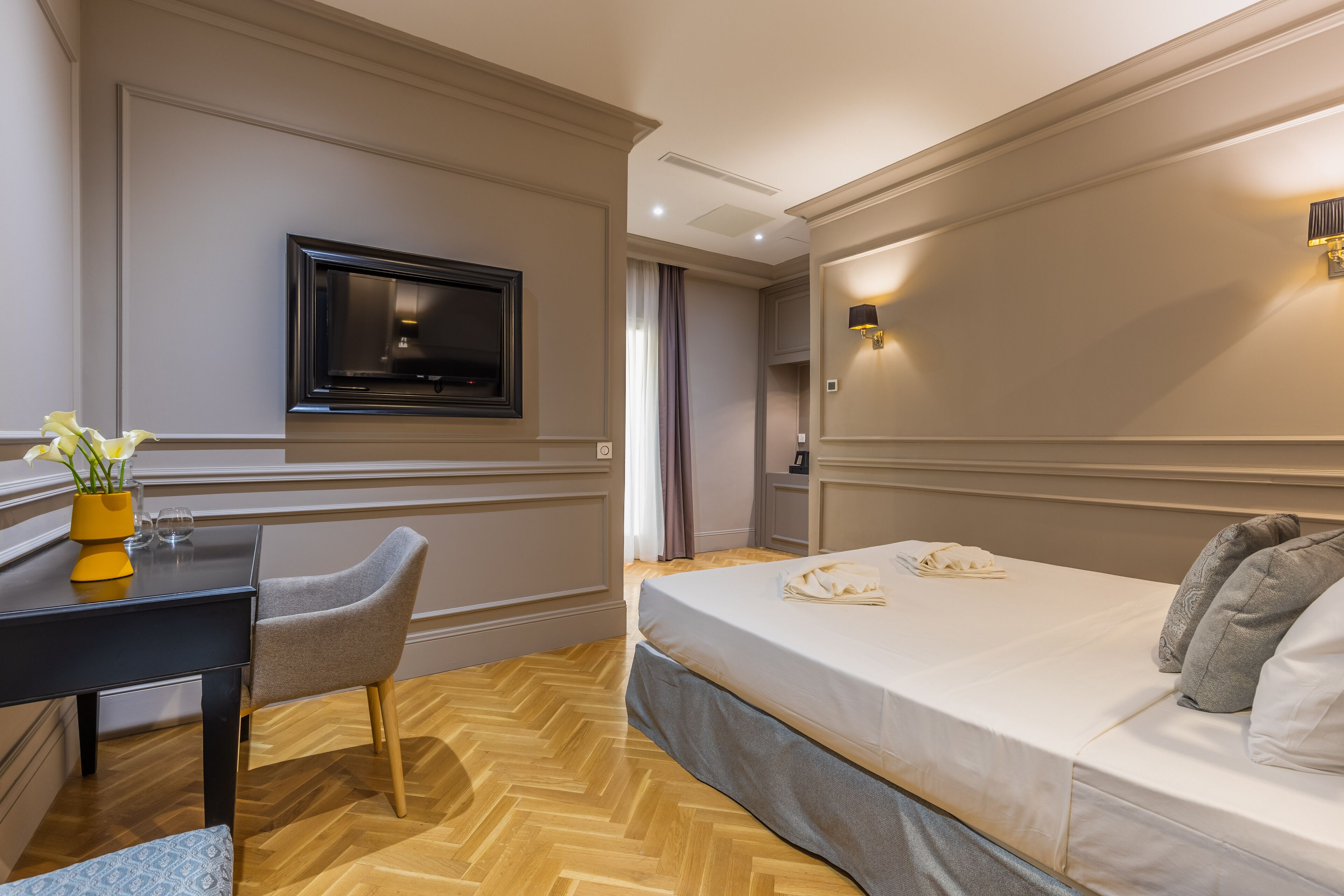 A hotel room with a bed, a desk, a chair, a TV, and a wall-mounted light.