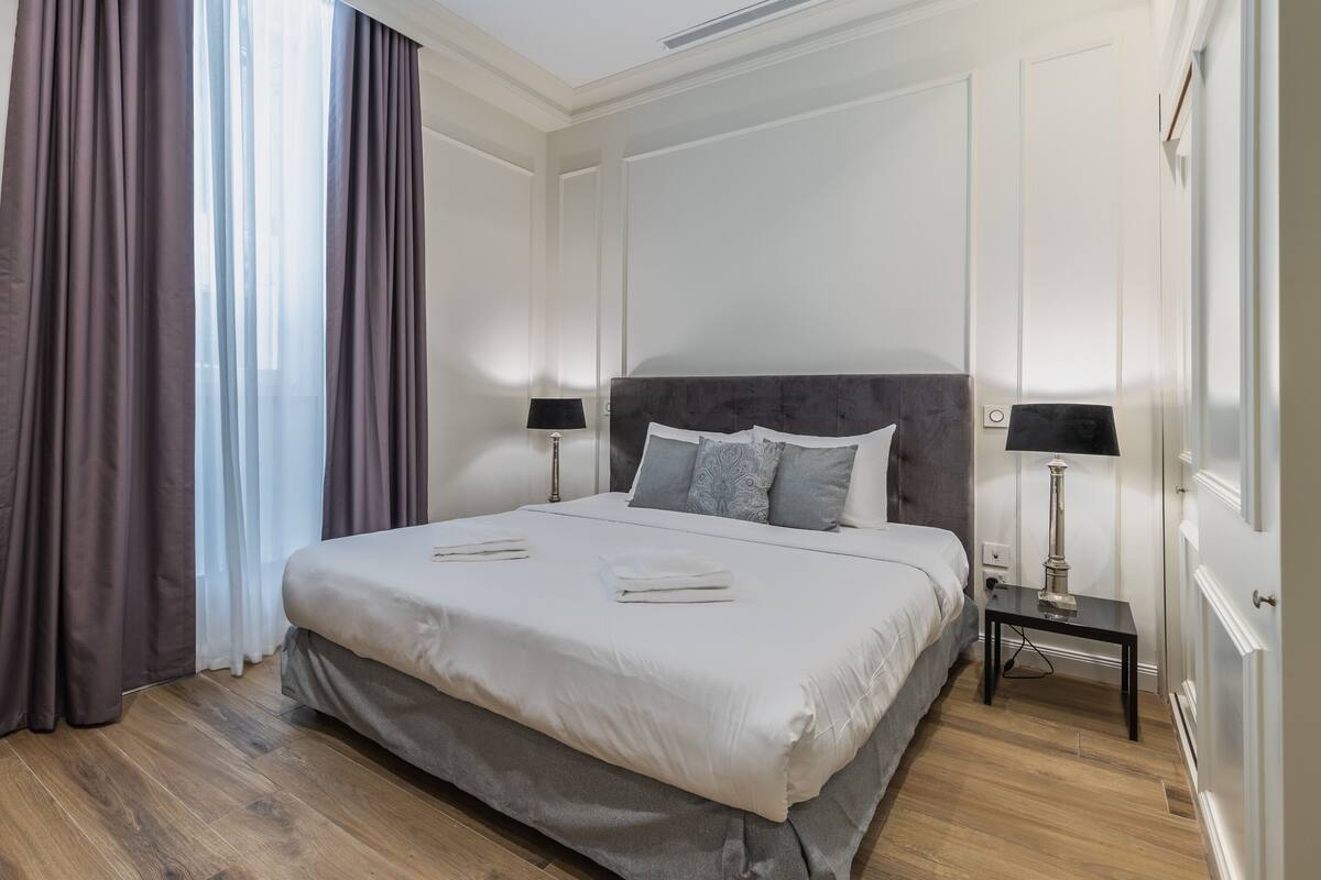 Una cama bien hecha con un cabecero gris, dos mesitas de noche con lámparas y un ventanal con cortinas.