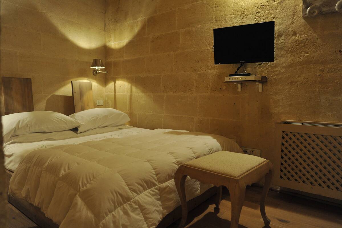 A hotel room with a bed, a wooden bench, a television mounted on the wall, and a radiator.