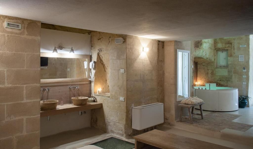 Un baño con paredes de piedra, un tocador de madera, dos lavamanos, un espejo y una pequeña mesa con un cojín.