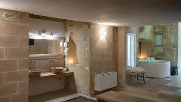 Un baño con paredes de piedra, un tocador de madera, dos lavamanos, un espejo y una pequeña mesa con un cojín.
