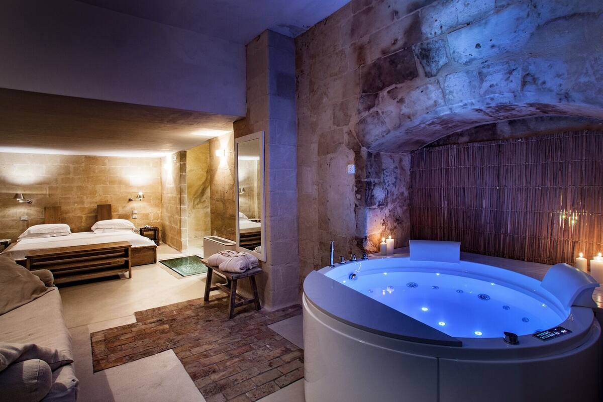 A modern spa room with a stone wall, a large bathtub, and a bed with a wooden headboard.