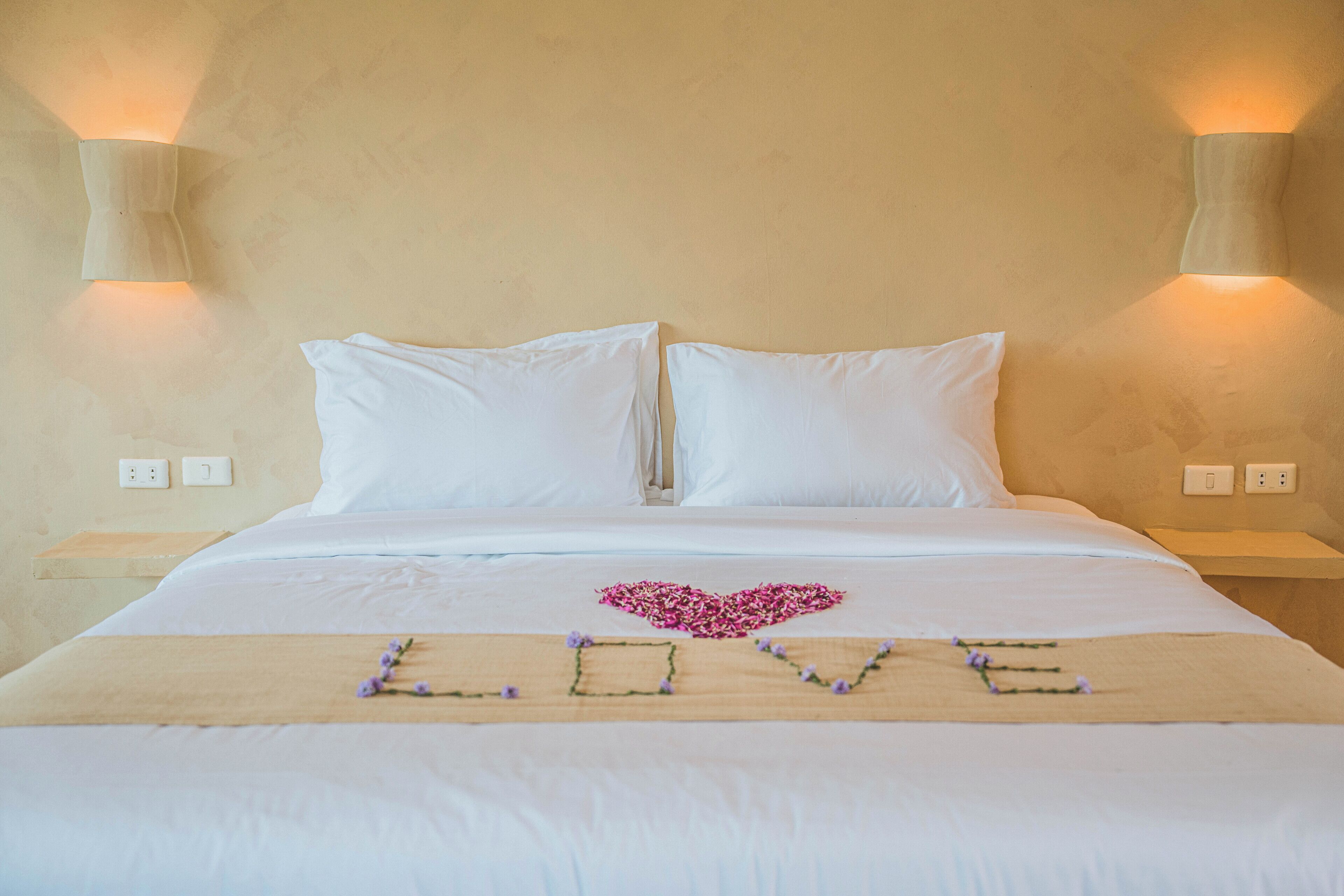Una cama bien hecha con sábanas blancas, una decoración con forma de corazón hecha de flores rosas, y la palabra 'LOVE' escrita con flores moradas sobre una corredera de madera.