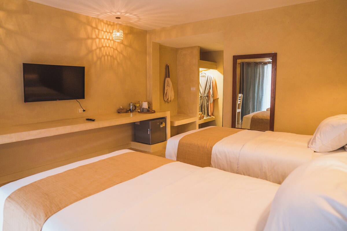 A hotel room with two beds, a TV mounted on the wall, a small refrigerator, and a mirror reflecting a window with curtains.