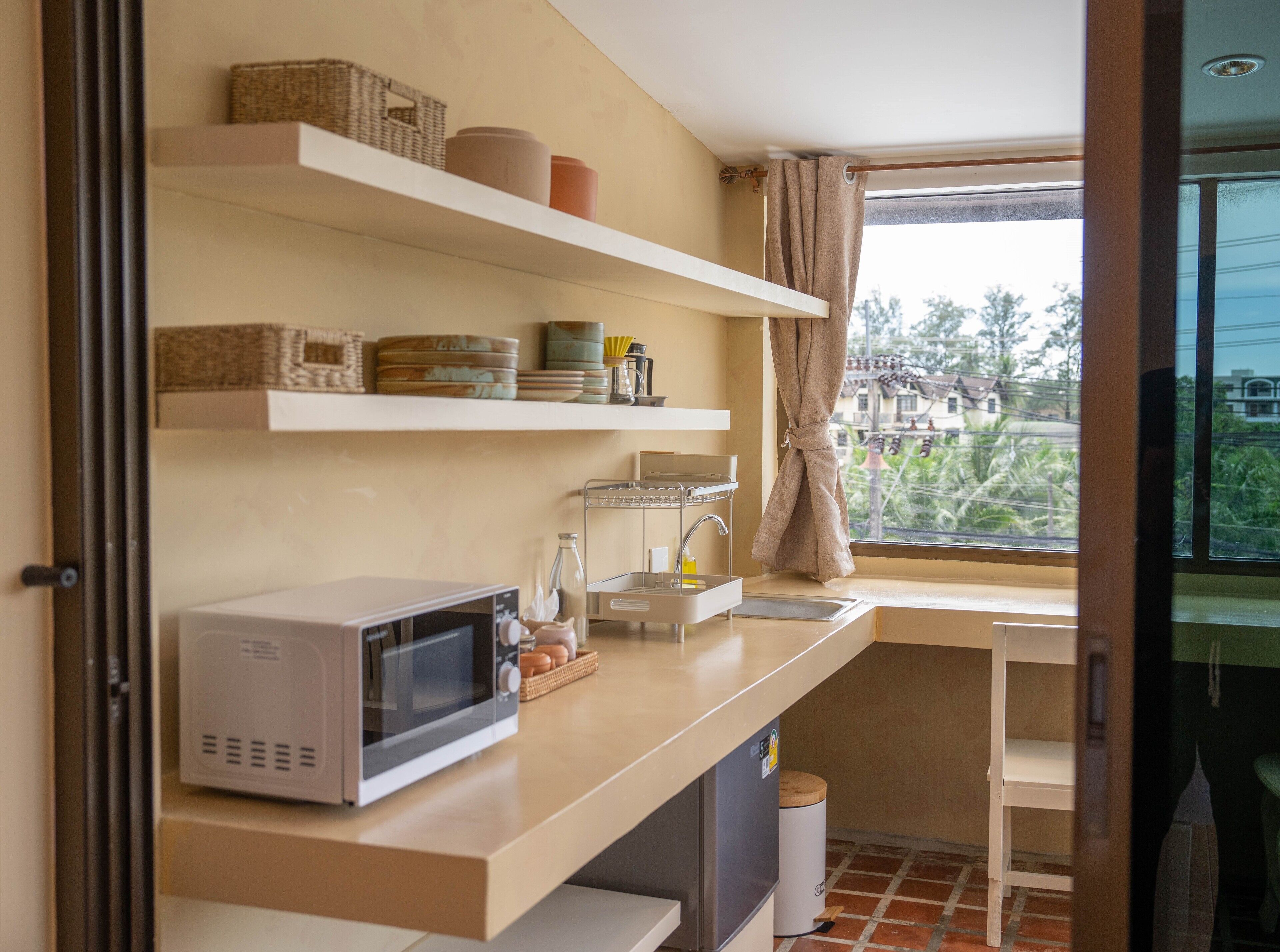 A kitchen with a microwave, shelves with kitchenware, a sink, and a window with a view of buildings and trees.