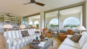 Amplia sala de estar con un sofá a rayas, sillones, una mesa de centro y una estantería. Grandes ventanas ofrecen vistas al exterior.
