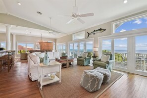 A spacious living room with a large window overlooking the ocean, a ceiling fan, and a dining area with a view. - Welcome to Land Yacht! (St. George Island)