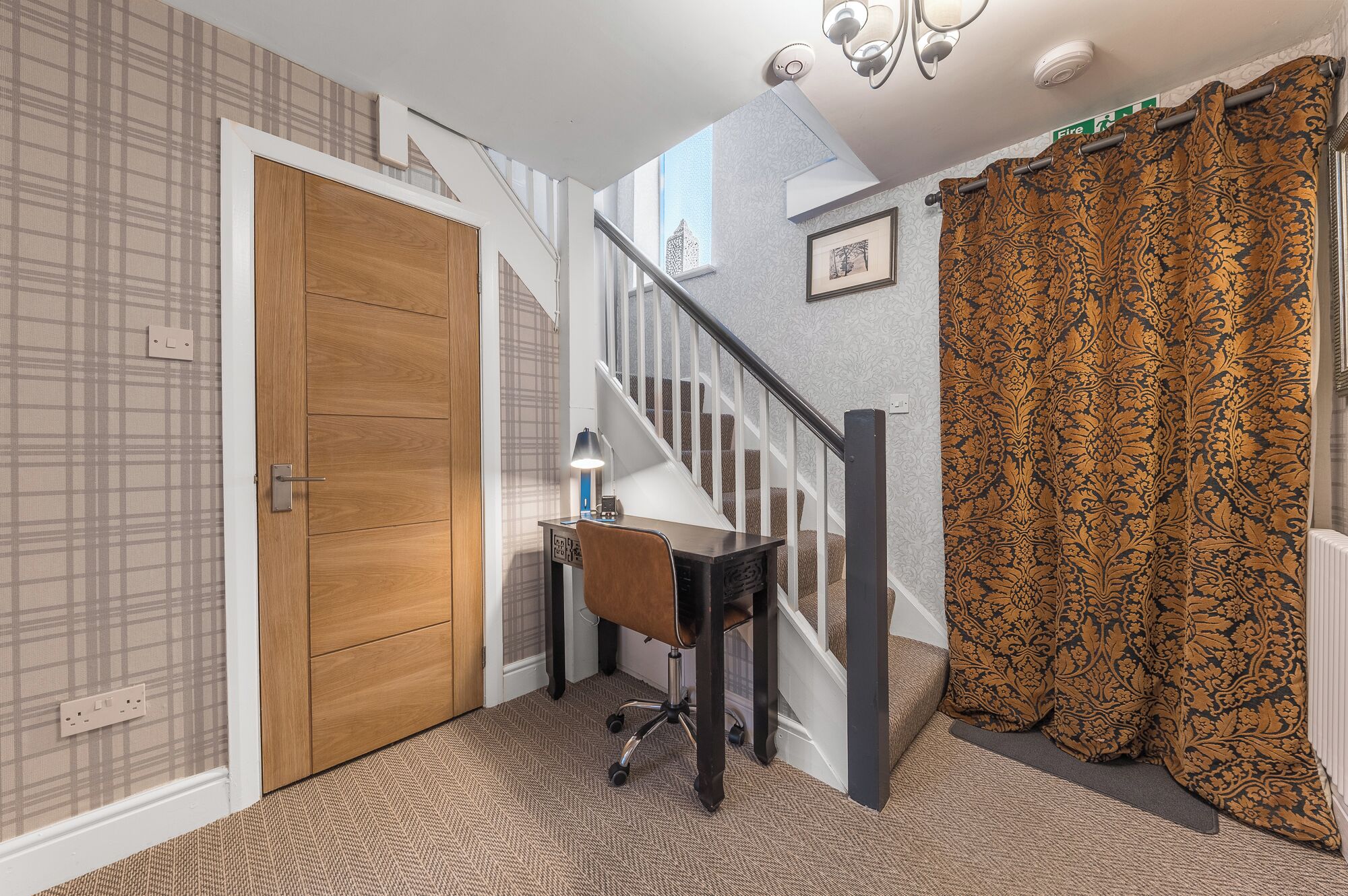 A hallway with a wooden door, a desk with a chair, a staircase, and patterned curtains.