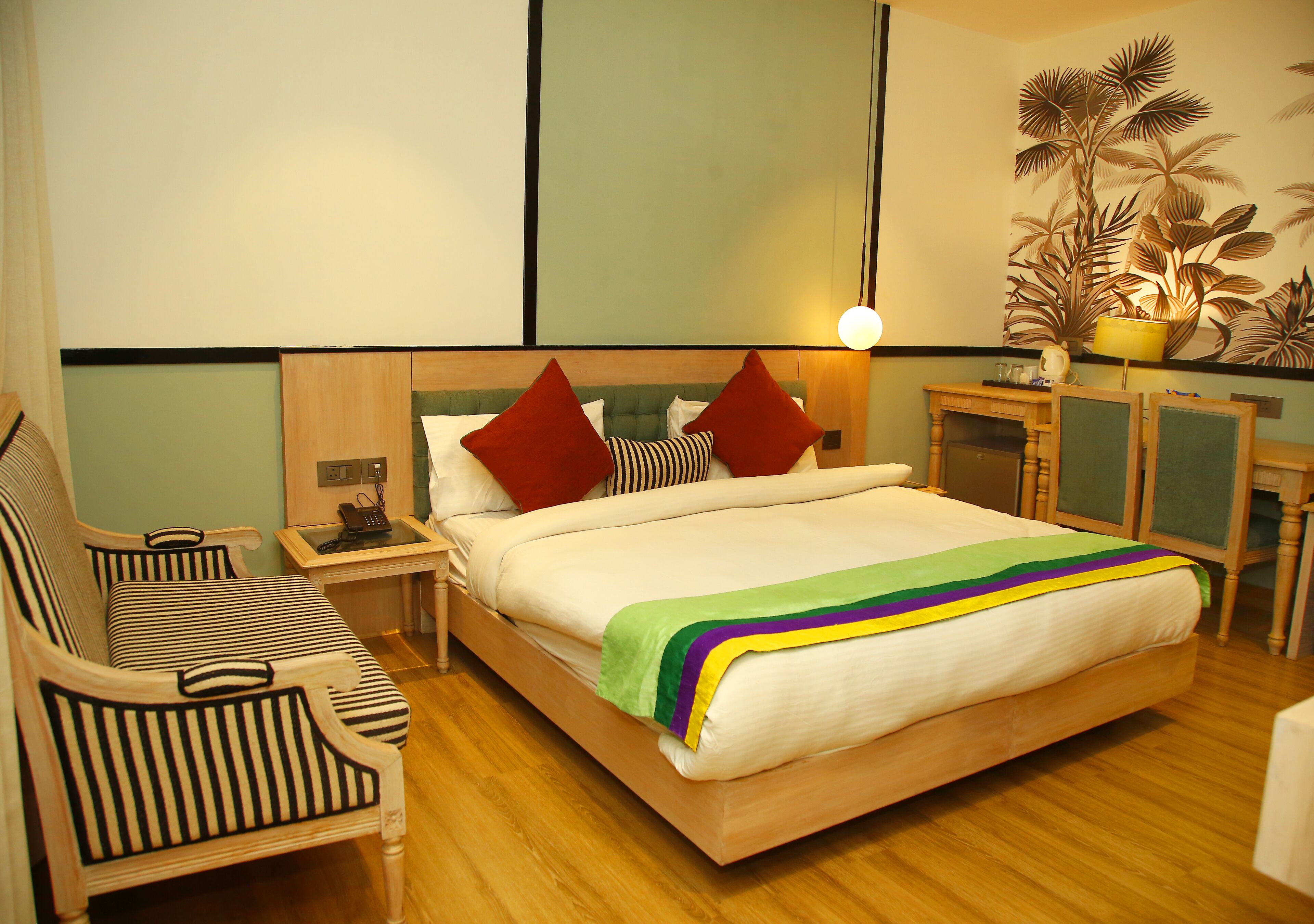 Habitación de hotel con una cama, dos sillas a rayas, una mesita de noche, una lámpara y una planta decorativa.