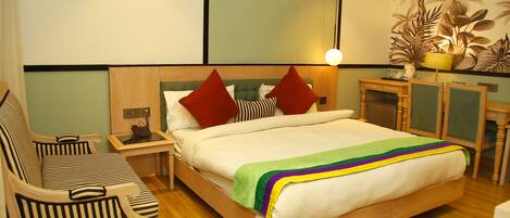 Habitación de hotel con una cama, dos sillas a rayas, una mesita de noche, una lámpara y una planta decorativa.