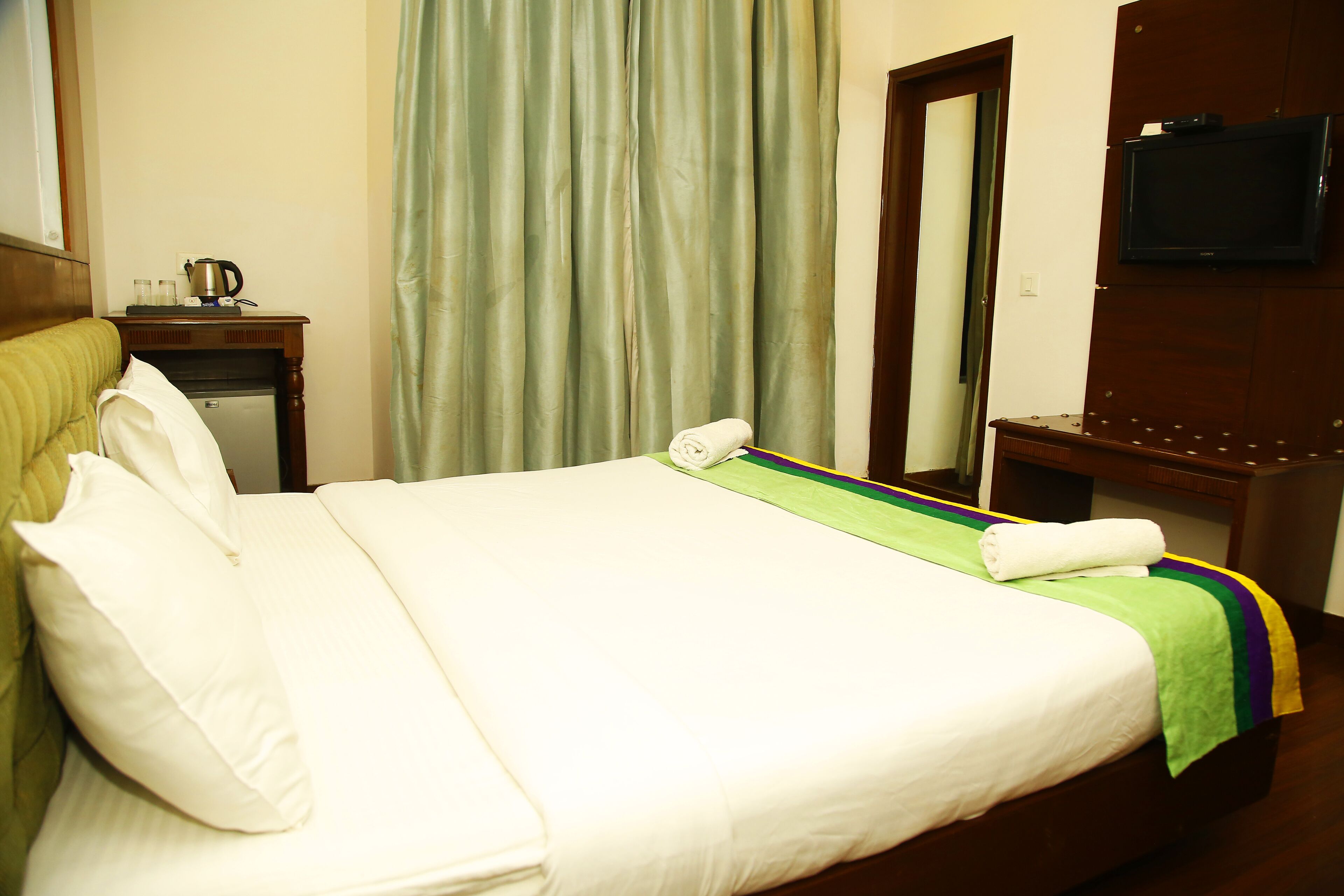 A hotel room with a neatly made bed, a television mounted on the wall, and a small table with a kettle.