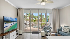 Una sala de estar con televisor, sofá, sillón y balcón con vistas a palmeras y una playa.
