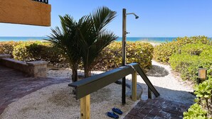 Een terras aan het strand met een houten bankje, een kleine palmboom en een pad naar het strand.