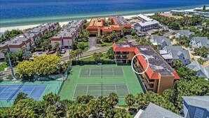 Luchtbeeld van een resort met meerdere gebouwen, een tennisbaan en een strand op de achtergrond.
