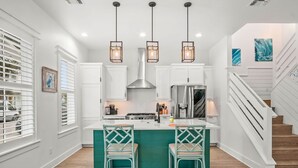A modern kitchen with white cabinetry, a central island with teal barstools, and pendant lights. - Resort Pools, Golf Cart Boardwalk to the Beach (Port Aransas)