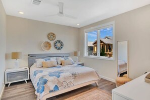 Une chambre à coucher avec un lit, des tables de chevet, un ventilateur de plafond et une fenêtre donnant sur une maison.