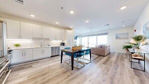 Una cocina moderna con armarios blancos, un comedor con sillas azules y un amplio ventanal que proporciona luz natural.