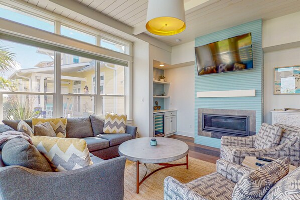 A spacious living room with a large sofa, a coffee table, and a fireplace. There are multiple windows providing natural light and a view of the outside.
