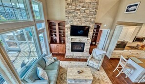 A spacious living area with a stone fireplace, a television, and a kitchen area with white cabinets.