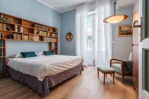 A bedroom with a large bed, a wooden bookshelf, a small chair, and a window with curtains. - Riva House Catania by Wonderful Italy (Catania)