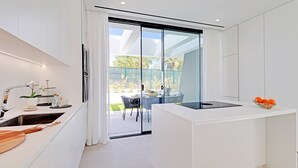 Une cuisine moderne dotée d’un îlot central, d’un coin repas avec une table et des chaises, et d’une porte-fenêtre coulissante donnant sur une terrasse aménagée avec du mobilier d’extérieur.