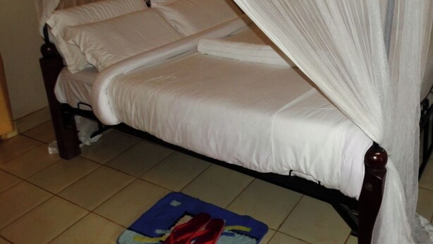 Een bed met hemelbed, wit beddengoed en een blauwe mat met rode schoenen op de vloer.