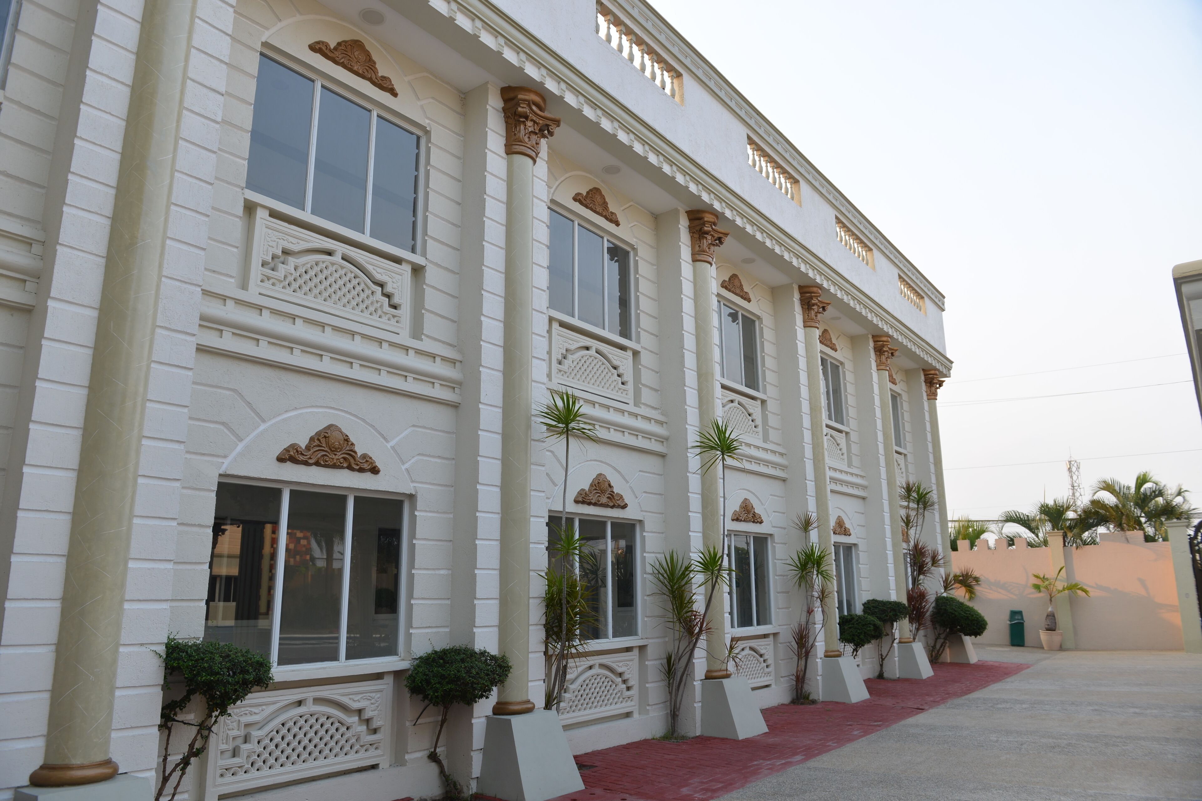 A large, white building with multiple windows and columns, surrounded by potted plants and a red pathway.