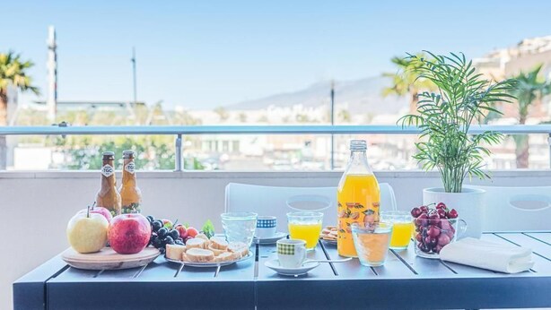 Una mesa de desayuno con frutas, bebidas y pan en un balcón con vistas a edificios y montañas.