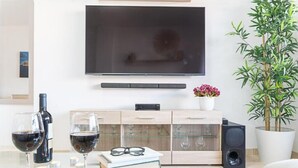 Una sala de estar moderna con un televisor de pantalla plana montado en la pared, un mueble de madera con puertas de cristal, una planta grande en maceta, y una mesa de vidrio con copas de vino y un libro.