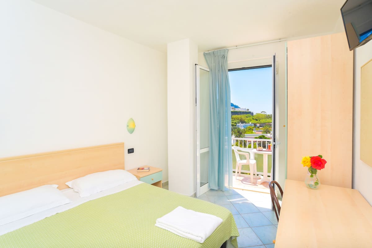 Ein Hotelzimmer mit Bett, Nachttisch, Tisch mit Blumenvase und Balkon mit Blick.