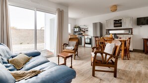 Ein Wohnzimmer mit blauem Sofa, Holzstühlen, einem kleinen Tisch und einer Küchenzeile mit Schränken und einem Fernseher.