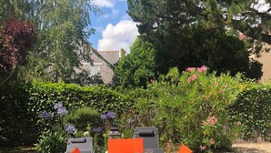Un área de comedor al aire libre con una larga mesa preparada para seis personas, rodeada de sillas anaranjadas, y un jardín y árboles al fondo.