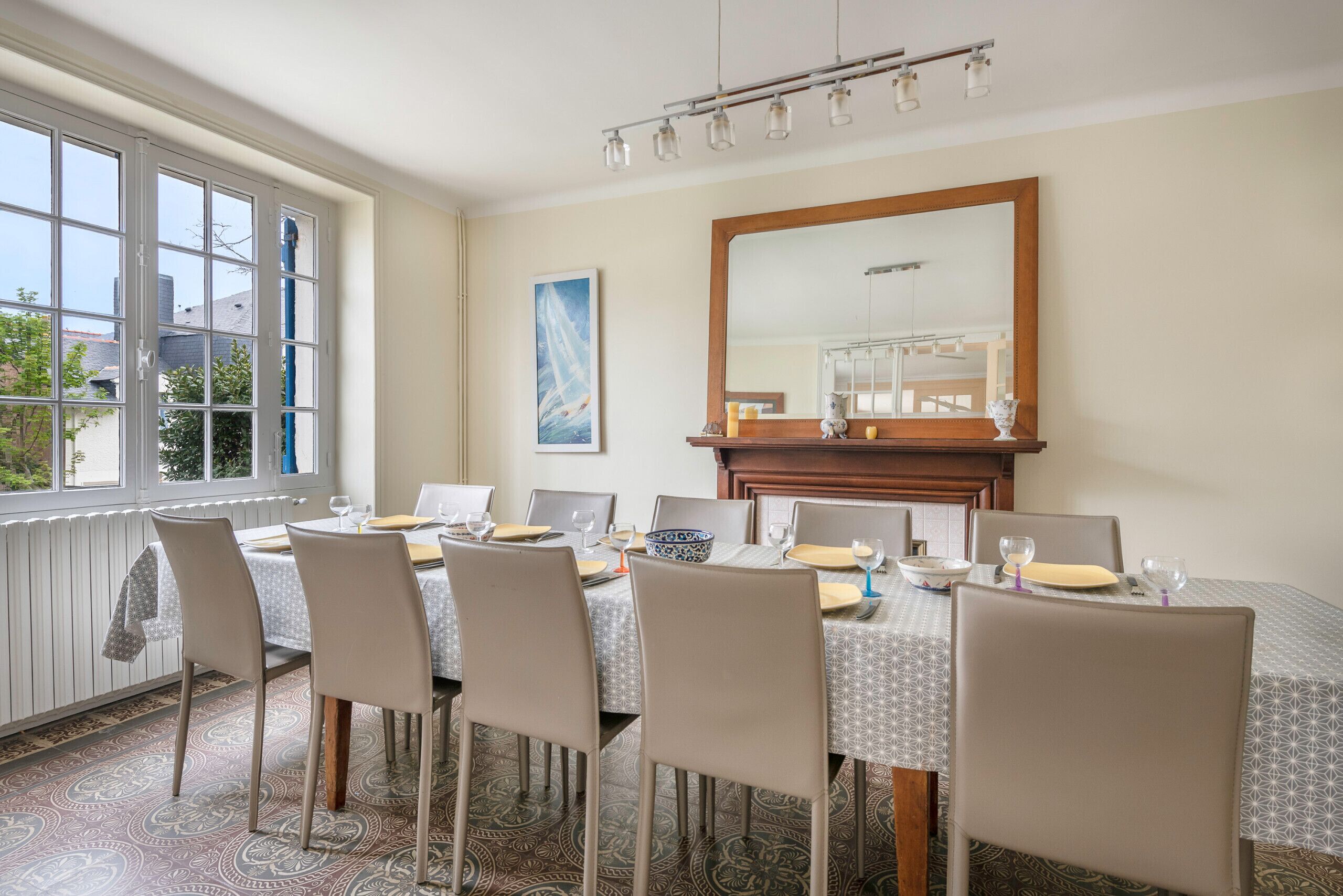 Une salle à manger avec une table dressée pour six personnes, un grand miroir au-dessus de la table et une fenêtre avec des stores.