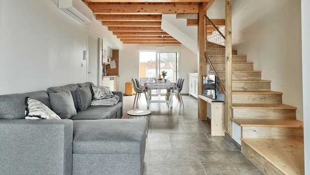 A modern living room with a grey sofa, wooden staircase, and a dining area with a table and chairs.