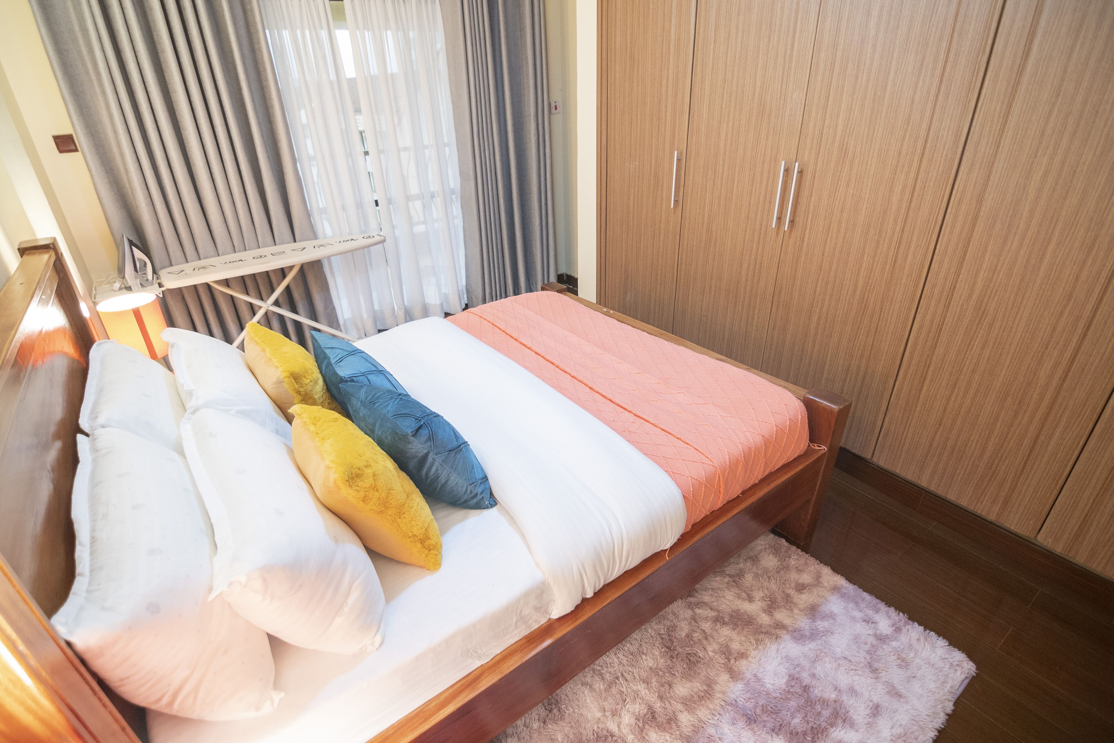 A bedroom with a wooden bed, white and yellow pillows, a pink blanket, a wooden wardrobe, and a window with curtains.