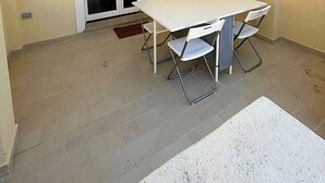 Un pequeño patio con una mesa y sillas blancas, una puerta corrediza de vidrio y piso de baldosas.