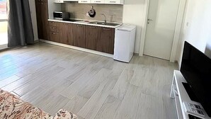 Cocina moderna con armarios blancos, microondas y un lavamanos. Se ve una puerta a la derecha y un televisor de pantalla plana sobre un mueble bajo.