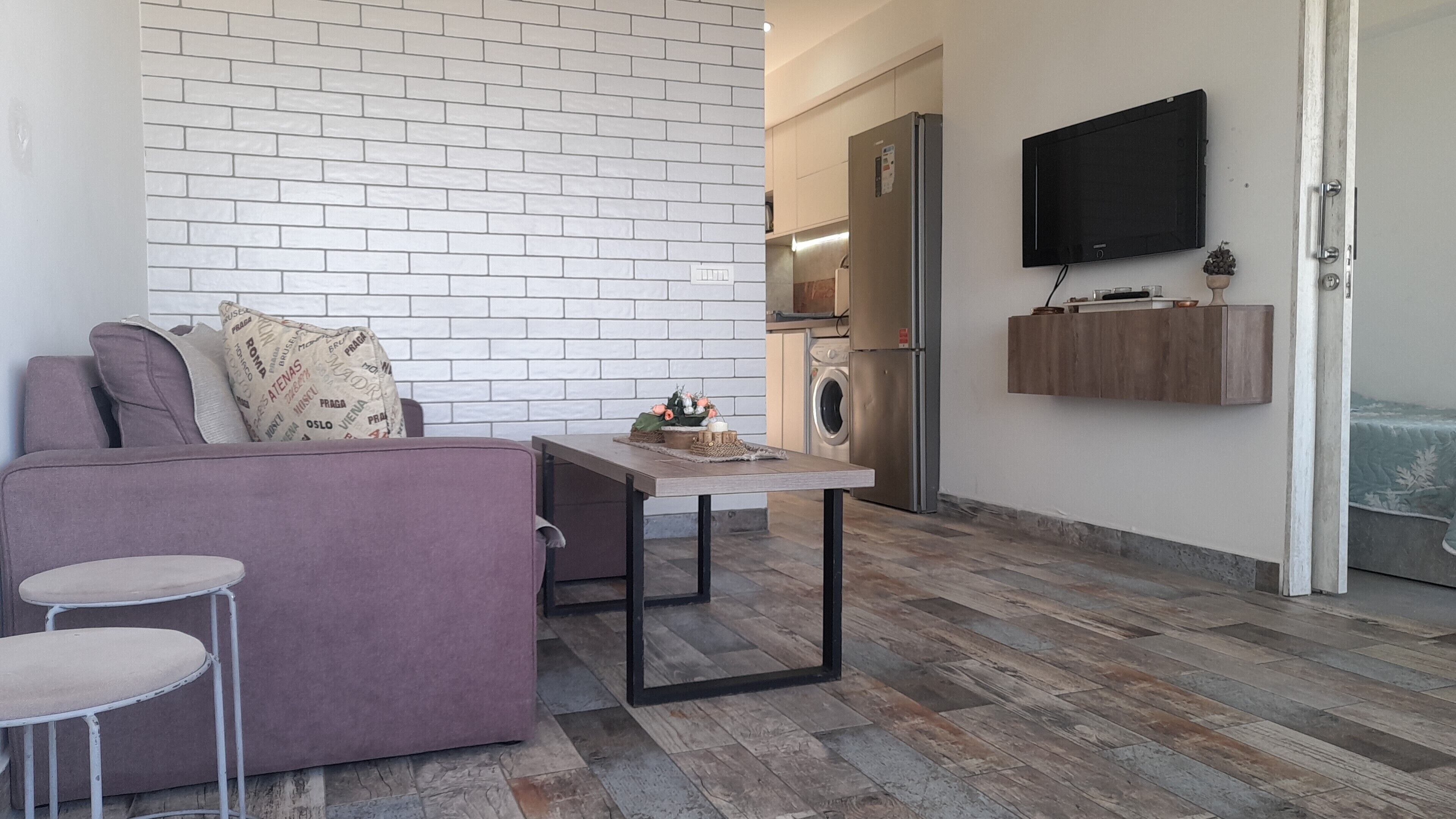 A modern apartment with a living area featuring a sofa, a small dining table, and a kitchen with a refrigerator and a washing machine.