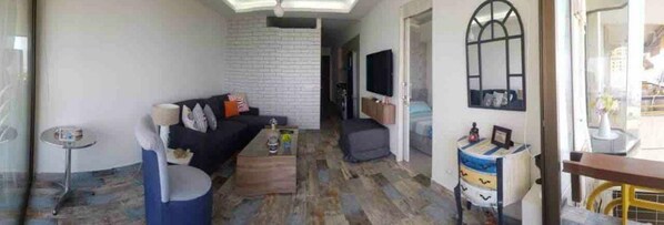 A living room with a sofa, coffee table, and a TV. There is a small table with a lamp and a chair. The room has a tiled floor and a window with a view. - 2 Bed Flat - Sleeps 6 - Pets - Pool - Parking (Jounieh)