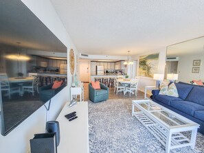A modern living room with a blue sofa, white coffee table, and a dining area with a white table and chairs.