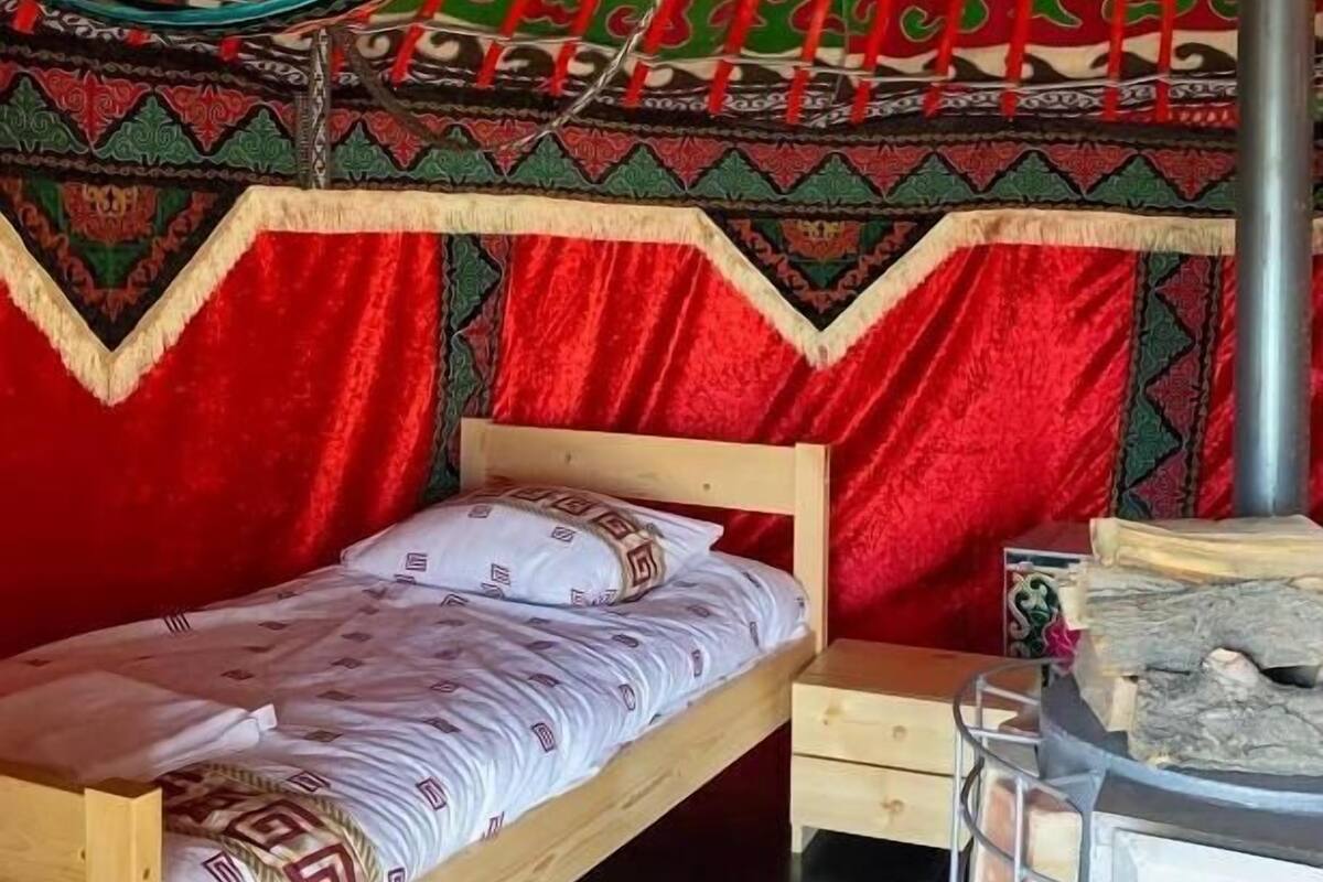 Een enkel bed met een bedsprei met patroon, een klein nachtkastje en een verwarming. De kamer heeft een wand met een rood-groen patroon en een rode hemelbed.