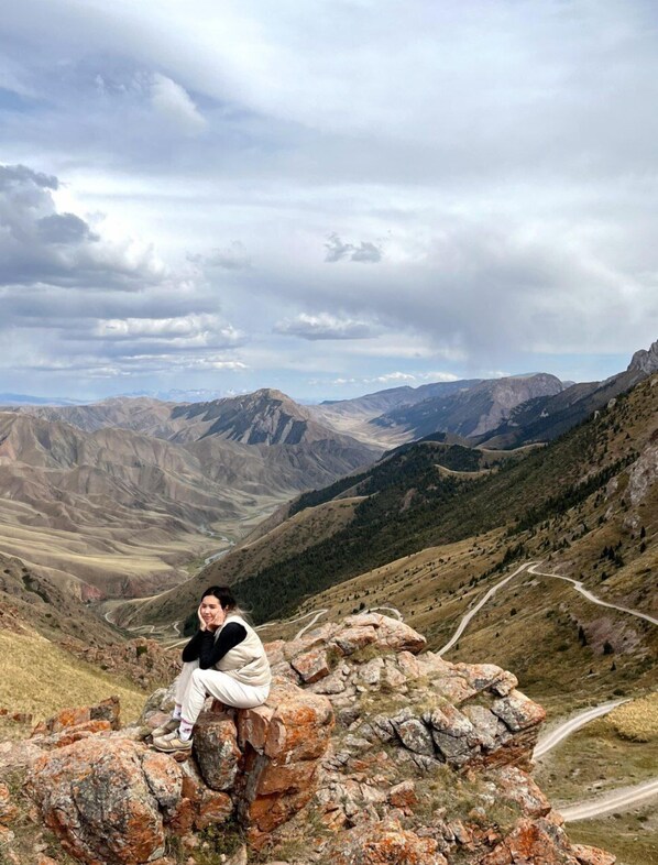 A person sitting on a rocky outcrop with a mountainous landscape in the background.