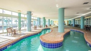 An indoor swimming pool with a curved design, surrounded by lounge chairs and tables, and large windows providing natural light. - Budget Friendly Condo for 2 in Myrtle Beach, Walk to the Waves! (Myrtle Beach)