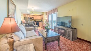 A living room with a sofa, coffee table, and TV, a kitchen in the background, and a patterned carpet. - Budget Friendly Condo for 2 in Myrtle Beach, Walk to the Waves! (Myrtle Beach)