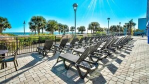 A sunlit outdoor seating area with lounge chairs and tables, a clear view of the ocean, and a paved walkway. - Budget Friendly Condo for 2 in Myrtle Beach, Walk to the Waves! (Myrtle Beach)