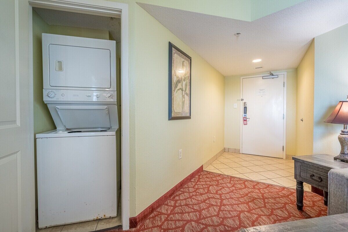 A laundry room with stacked washers and dryers, a door leading to another room, a patterned rug, and a small table with a lamp.