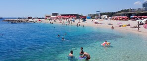 Une plage avec une eau claire, des gens qui se baignent et qui prennent des bains de soleil, et des parasols de plage.