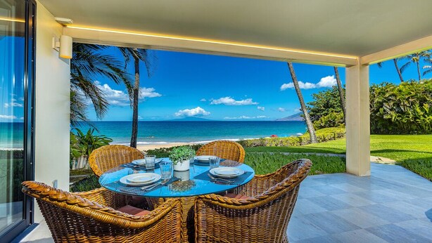 A dining area with wicker chairs and a glass table overlooking a beach with palm trees and the ocean. - Polo Beach Club 106 | Beachfront Wailea Condo with Direct Beach Access (Wailea)